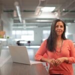 Smiling woman in a bright office, standing by her laptop, exuding confidence.